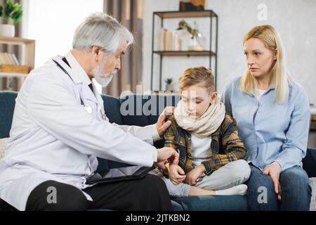 Front view of good-looking professional senior male caucasian doctor in medical coat, consulting his teen boy patient wihis mom, touching hand of child to check pulse, during home visit. Stock Photo