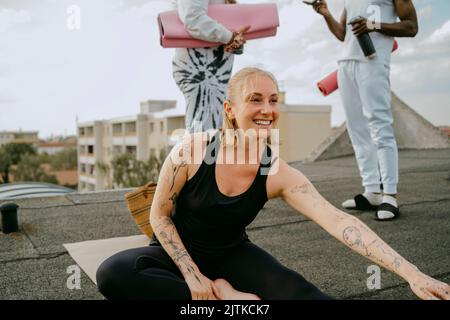 Happy woman practicing yoga while friends talking with each other standing on rooftop Stock Photo