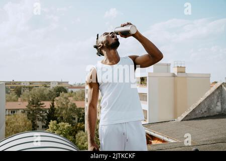 Young man drinking water from bottle while standing on rooftop during sunny day Stock Photo