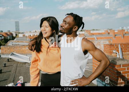 Smiling young man standing with arm around female friend looking away while standing on rooftop Stock Photo