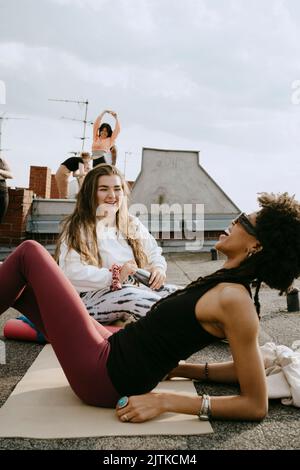 Smiling young woman talking with cheerful female friend reclining on exercise mat Stock Photo