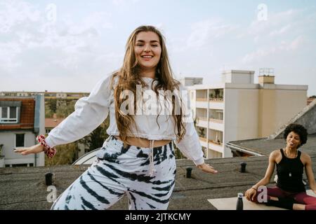 Portrait of smiling young woman practicing yoga by friend on rooftop Stock Photo