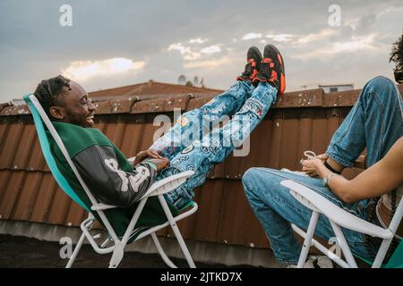 Side view of cheerful young man talking with female friend while sitting on chair at rooftop Stock Photo