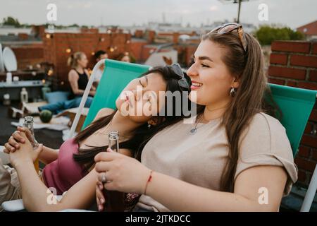 Young woman with eyes closed leaning on female friend holding beer bottle Stock Photo