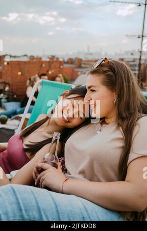 Smiling young woman holding beer bottle sitting by female friends with eyes closed on rooftop Stock Photo