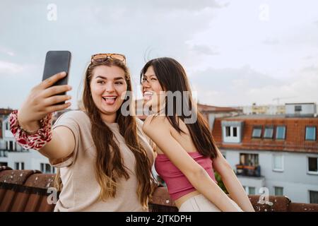 Happy women sticking out tongue while taking selfie through smart phone on rooftop Stock Photo