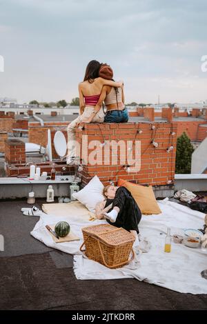 Woman lying on blanket with female friends sitting with arms around at rooftop Stock Photo