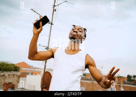 Portrait of young man holding smart phone while gesturing peace sign Stock Photo