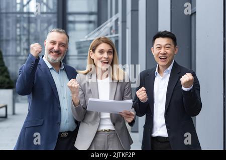 Businesswoman boss with her diverse team looking at the camera and rejoicing in success and victory, celebrating a successful contract a team of colleagues outside the office building with documents. Stock Photo
