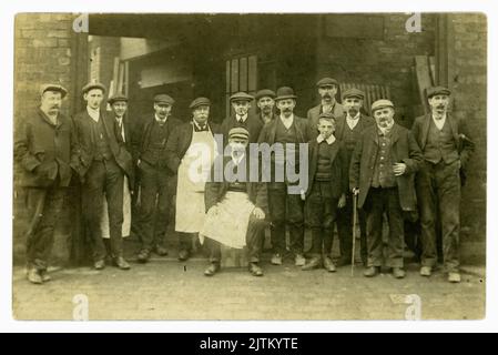 Original clear postcard of working class men, factory workers & young apprentice boy. Some are wearing aprons, all wear hats - flat caps and a bowler hat, and suits, boots with steel toe caps, lots pf characters, dated November 1912 on reverse, UK. Stock Photo