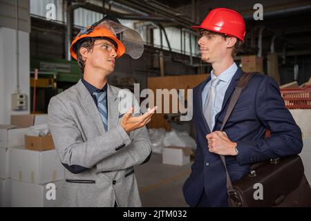 Two colleagues during a conversation at work in the factory, engineers elegantly dressed wear protective helmet Stock Photo