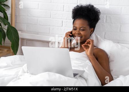 Happy attractive african american girl working office work remotely, Black woman using laptop computer and phone sitting in bed at home, Distance Stock Photo
