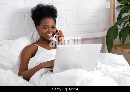 Happy attractive african american girl working office work remotely, Black woman using laptop computer and phone sitting in bed at home, Distance Stock Photo