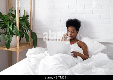 Happy attractive african american girl working office work remotely, Black woman using laptop computer and phone sitting in bed at home, Distance Stock Photo