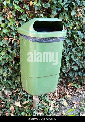 Vertical shot on a green plastic public wastebasket for disposal of garbage against a wall with green ivy Stock Photo