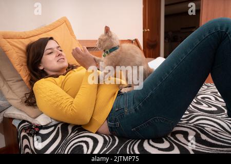 Beautiful Hispanic young woman smiling dressed in yellow jacket, lying on her bed playing with her little kitten with her hands Stock Photo