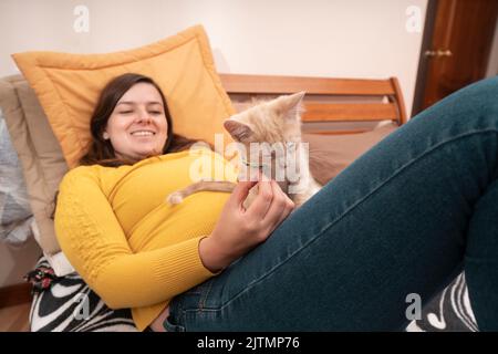 Beautiful Hispanic young woman smiling dressed in yellow jacket, lying on her bed playing with her little kitten. focused cat Stock Photo