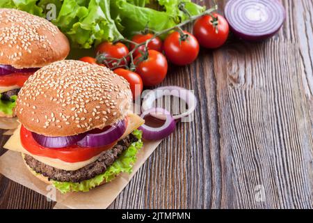 Two homemade cheeseburgers with beef patties , fresh salad, tomatoes and onion on seasame buns, served on brown wooden table. Stock Photo