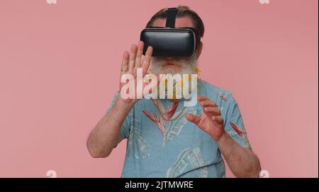 Exited elderly stylish man use virtual reality futuristic technology VR app headset helmet to play simulation 3D 360 video game, drawing. Grandfather on pink studio background. People sincere emotions Stock Photo