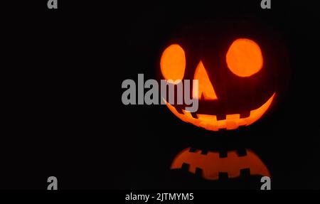 Close-up on a Jack O' Lantern lit in the dark and reflecting on a black surface in front of it. Stock Photo