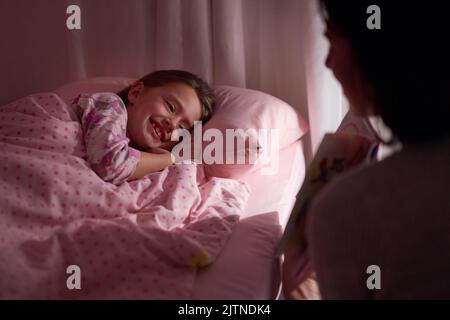 Dont forget to show me the pictures Mom. a little girl lying in bed while her mom reads a bedtime story.