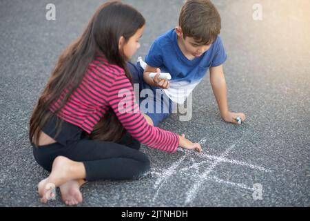 Is it my turn yet. a cute little boy playing tic tac toe with his sister on the pavement outside their house. Stock Photo