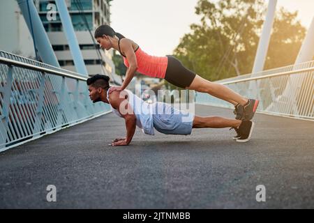 Behind every strong man is a strong woman. a young woman balancing on her boyfriends back while doing pushups outdoors on a bridge. Stock Photo