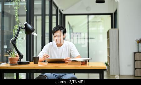Handsome young Asian male freelance graphic designer or office worker in casual outfit using digital tablet at his office desk. Stock Photo