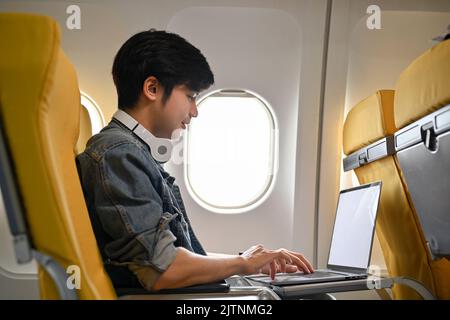 Hipster millennial Asian male traveler passenger using laptop computer to manage and clear his task during the flight. Urban lifestyle and remote work Stock Photo