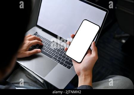 close-up image, A businessman is on a flight for his business trip, using his portable laptop and smartphone on the plane. phone white screen mockup. Stock Photo