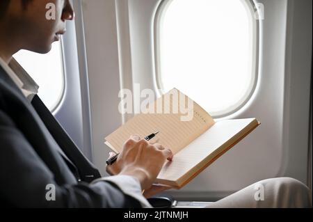 Smart and professional young Asian businessman is on the flight, reading, checking his schedule or writing something on his notebook or diary. cropped Stock Photo