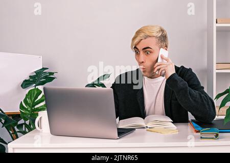 A young office worker is shocked and surprised looking at a laptop talking on the phone, sitting at a desk in an office surrounded by documents Stock Photo