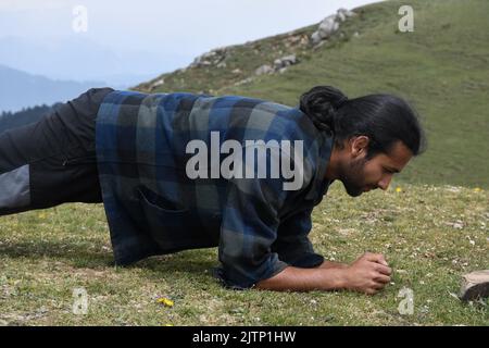 Side view of a handsome young Indian man with ponytail hair doing plank exercise in the mountain Stock Photo