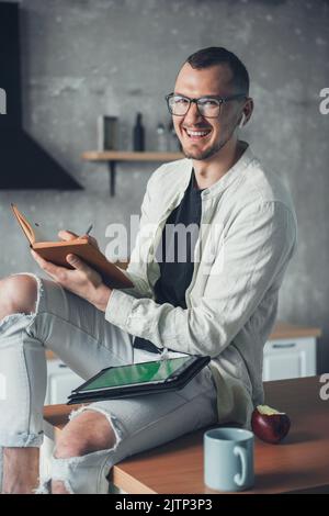 Caucasian man having coffee in the morning at kitchen working with a tablet PC and writing in an open notepad. Distance education. Education business Stock Photo