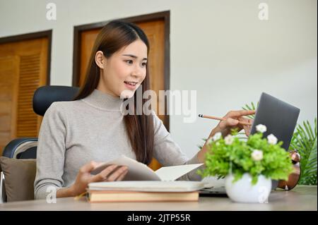 Charming and pretty young Asian female college student doing her project assignment at home, using laptop to search the online information. Stock Photo