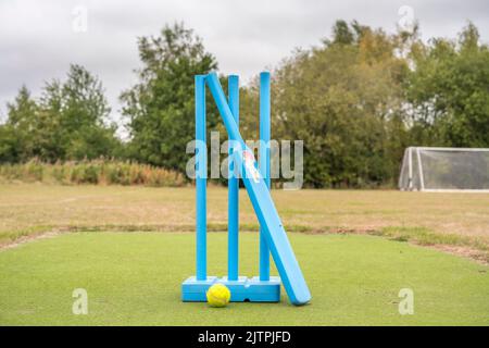 Close up of blue cricket stumps, blue cricket bat and yellow tennis ball set up outdoors ready for a children's junior version of the game. Stock Photo