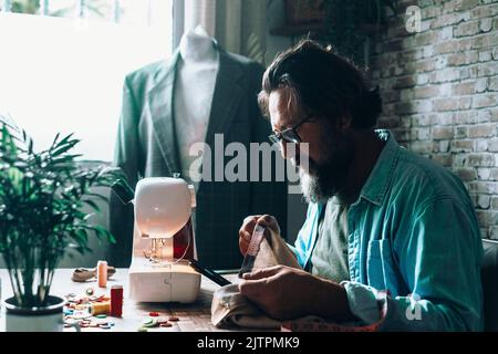 Man at home using sewing machine to tailor and modify garments. Blazer in background. Tailoring and tailor at work at the workshop. Concept of people Stock Photo