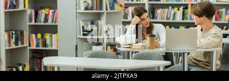 smiling teen girl talking to friend near laptop and books in library, banner,stock image Stock Photo