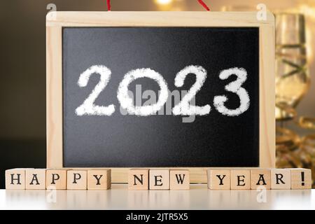 Blackboard with 2023 chalk text and happy new year made of wooden blocks witg festive champagne in the background. New Year's Eve concept Stock Photo
