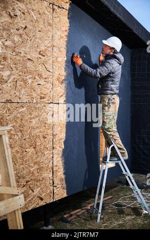 Male painter using paint roller, doing exterior paint work in a black color, standing on a ladder. Man worker building wooden frame house. Carpentry and construction concept. Stock Photo
