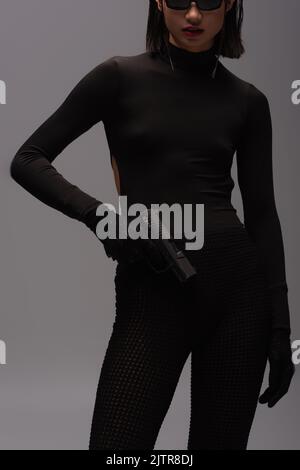 cropped view of dangerous asian woman in total black outfit and stylish sunglasses holding gun isolated on grey,stock image Stock Photo