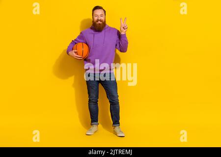 Full length photo of funky cute man wear violet sweatshirt showing v-sign holding ball isolated yellow color background Stock Photo