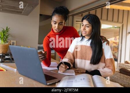 Biracial mother pointing at laptop while assisting daughter in writing homework at home Stock Photo