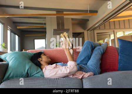 Biracial teenage girl reading book while lying on couch in living room Stock Photo