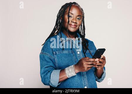 Beautiful black woman looking at the camera while holding a smartphone and standing against a grey background. Mature woman with dreadlocks wearing a