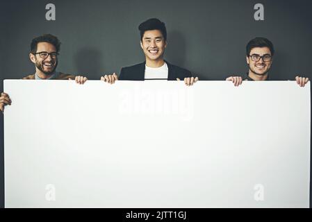 A group of young, smiling and professional business men holding a white sign copyspace portrait. A team of male designers standing together with a Stock Photo