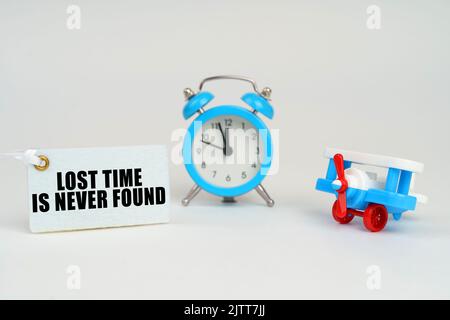 On a white surface there is a toy plane, an alarm clock and a sign with the inscription - Lost Time is Never Found. Business concept. Stock Photo