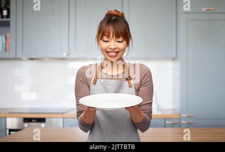 happy woman in apron with empty plate in kitchen Stock Photo