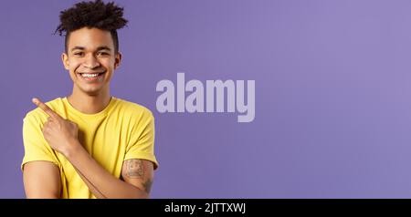 Close-up portrait of enthusiastic, happy young hipster male with dreads, beaming smile and pointing finger upper left corner, present cool product Stock Photo