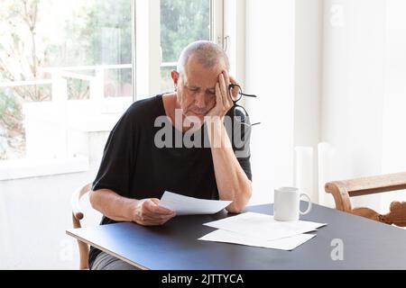 Frustrated man reading paper documents while sitting at table with glasses in hand holding his head. Stock Photo